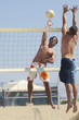 Young man jumping, hitting volleyball over net on beach; opponent defending