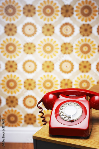 Foto: Old fashioned red telephone on table in front of flowery wallpaper Old fashioned red telephone on table in front of flowery wallpaper