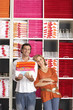 Photo: Couple shopping in department store, woman leaning against boyfriend, man holding pile of folded towels, smiling, front view, portrait, shelves in background Couple shopping in department store, woman leaning against boyfriend, man holding pile of folded towels, smiling, front view, portrait, shelves in background
