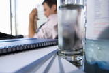 Photo: Glass of water by paperwork on desk, young businessman in background (differential focus) Glass of water by paperwork on desk, young businessman in background (differential focus)