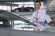 Photo: Car salesman standing beside new saloon cars in showroom, holding brochure, smiling, portrait Car salesman standing beside new saloon cars in showroom, holding brochure, smiling, portrait
