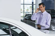 Photo: Car salesman standing beside new car in showroom, holding brochure, using mobile phone, smiling Car salesman standing beside new car in showroom, holding brochure, using mobile phone, smiling
