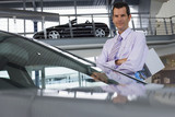 Photo: Car salesman standing beside new saloon cars in showroom, holding brochure, smiling, portrait Car salesman standing beside new saloon cars in showroom, holding brochure, smiling, portrait