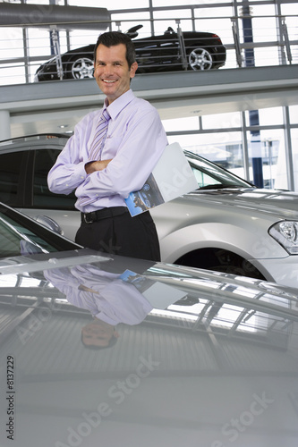 Photo: Car salesman standing beside new saloon cars in showroom, holding brochure, smiling, portrait Car salesman standing beside new saloon cars in showroom, holding brochure, smiling, portrait