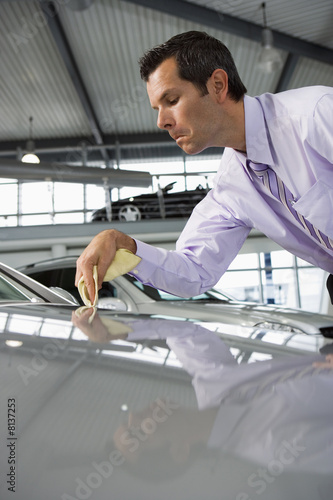 Photo: Car salesman polishing bonnet of new silver car with cloth in showroom (surface level) Car salesman polishing bonnet of new silver car with cloth in showroom (surface level)