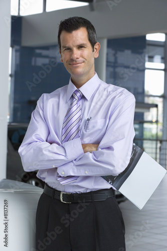 Photo: Car salesman standing in showroom, holding brochure, arms folded, smiling, portrait Car salesman standing in showroom, holding brochure, arms folded, smiling, portrait