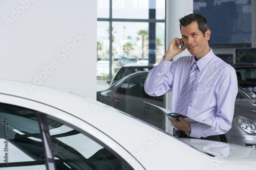 Photo: Car salesman standing beside new car in showroom, holding brochure, using mobile phone, smiling Car salesman standing beside new car in showroom, holding brochure, using mobile phone, smiling
