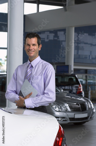 Photo: Car salesman standing beside new car in showroom, holding brochure, smiling, portrait Car salesman standing beside new car in showroom, holding brochure, smiling, portrait