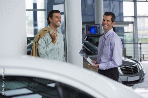 Photo: Car salesman standing with male customer in car showroom, holding brochure, smiling, side view, portrait Car salesman standing with male customer in car showroom, holding brochure, smiling, side view, portrait