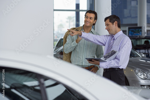 Photo: Car salesman pointing out new cars to male customer in showroom, holding brochure, smiling Car salesman pointing out new cars to male customer in showroom, holding brochure, smiling