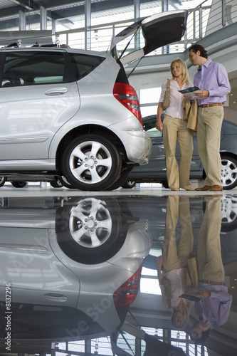 Photo: Couple looking at new silver hatchback in car showroom, man holding brochure, side view (surface level) Couple looking at new silver hatchback in car showroom, man holding brochure, side view (surface level)