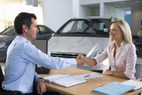 Photo: Car salesman and female customer sitting at desk in car showroom, shaking hands, smiling, side view Car salesman and female customer sitting at desk in car showroom, shaking hands, smiling, side view