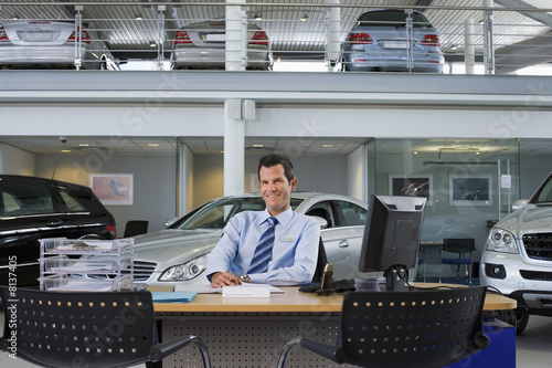 Photo: Car salesman sitting at desk in large car showroom, smiling, front view, portrait Car salesman sitting at desk in large car showroom, smiling, front view, portrait