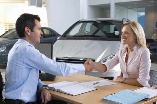 Photo: Car salesman and female customer sitting at desk in car showroom, shaking hands, smiling, side view Car salesman and female customer sitting at desk in car showroom, shaking hands, smiling, side view