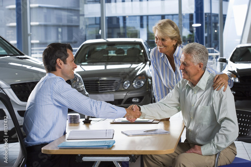 Photo: Car salesman and senior couple sitting at desk in large car showroom, two men shaking hands, smiling, side view Car salesman and senior couple sitting at desk in large car showroom, two men shaking hands, smiling, side view