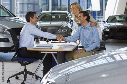 Photo: Car salesman and couple sitting at desk in large car showroom, two men shaking hands, smiling, side view Car salesman and couple sitting at desk in large car showroom, two men shaking hands, smiling, side view