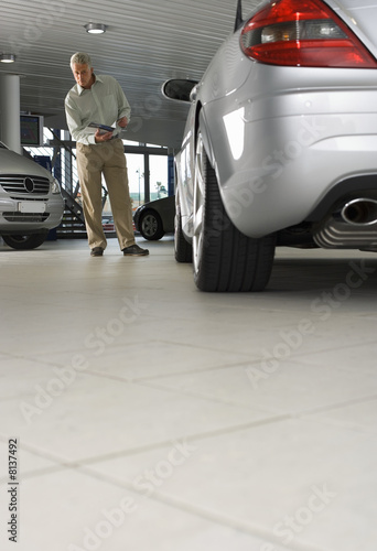 Photo: Senior man looking at new silver convertible car in large showroom, holding brochure (surface level) Senior man looking at new silver convertible car in large showroom, holding brochure (surface level)