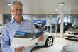 Photo: Senior man standing in large car showroom, looking at brochure, smiling, portrait Senior man standing in large car showroom, looking at brochure, smiling, portrait