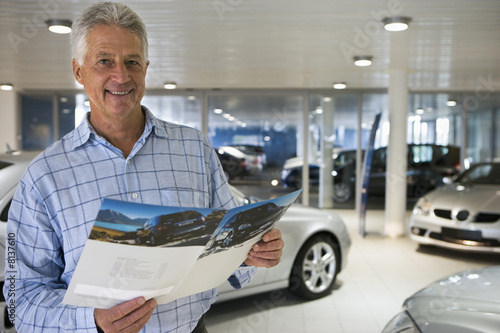Photo: Senior man standing in large car showroom, looking at brochure, smiling, portrait Senior man standing in large car showroom, looking at brochure, smiling, portrait