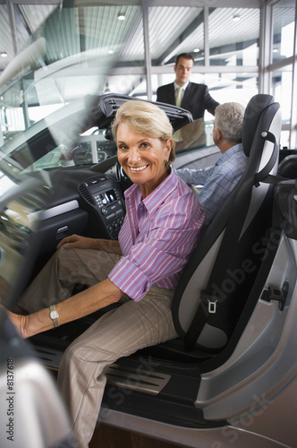 Photo: Couple looking at new convertible car in showroom, man talking to salesman, focus on woman sitting in passenger seat in foreground, smiling, side view, portrait Couple looking at new convertible car in showroom, man talking to salesman, focus on woman sitting in passenger seat in foreground, smiling, side view, portrait