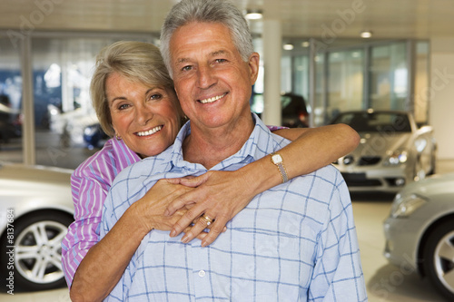 Photo: Senior couple standing in large car showroom, woman embracing man, smiling, front view, portrait Senior couple standing in large car showroom, woman embracing man, smiling, front view, portrait