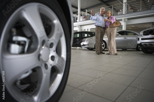 Photo: Senior couple standing in large car showroom, focus on wheel of new car in foreground (surface level) Senior couple standing in large car showroom, focus on wheel of new car in foreground (surface level)