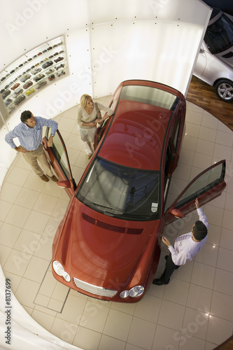 Photo: Car salesman showing couple new red hatchback in large car showroom, overhead view Car salesman showing couple new red hatchback in large car showroom, overhead view