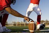 Photo: American football player attempting to kick field goal, teammate holding ball vertically against pitch (surface level) American football player attempting to kick field goal, teammate holding ball vertically against pitch (surface level)