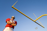 Photo: American football player, in red football strip and protective helmet, standing below goal post, arms folded, portrait, low angle view American football player, in red football strip and protective helmet, standing below goal post, arms folded, portrait, low angle view