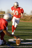 Photo: American football player attempting to kick field goal, teammate holding ball vertically against pitch American football player attempting to kick field goal, teammate holding ball vertically against pitch
