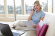Photo: Mature woman sitting on sofa with laptop, holding document, using mobile phone, smiling, side view Mature woman sitting on sofa with laptop, holding document, using mobile phone, smiling, side view