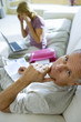 Photo: Mature woman sitting on sofa at home, using laptop and mobile phone, focus on senior man with bills in laptop, smiling, side view, portrait, elevated view Mature woman sitting on sofa at home, using laptop and mobile phone, focus on senior man with bills in laptop, smiling, side view, portrait, elevated view