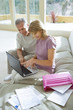 Photo: Senior couple sitting on sofa at home, using laptop, smiling, side view, pink folder and bills in foreground Senior couple sitting on sofa at home, using laptop, smiling, side view, pink folder and bills in foreground