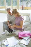 Photo: Senior couple sitting on sofa at home, using laptop, smiling, side view, pink folder and bills in foreground Senior couple sitting on sofa at home, using laptop, smiling, side view, pink folder and bills in foreground