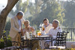 Photo: Two mature couples dining at outdoor restaurant table, standing couple placing hands affectionately on shoulders of seated friends, smiling Two mature couples dining at outdoor restaurant table, standing couple placing hands affectionately on shoulders of seated friends, smiling