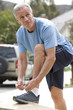 Photo: Active senior man, in blue t-shirt and shorts, tying trainer shoelace on driveway wall, side view Active senior man, in blue t-shirt and shorts, tying trainer shoelace on driveway wall, side view