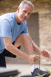 Photo: Active senior man, in blue t-shirt and shorts, tying trainer shoelace on driveway wall, smiling, side view, portrait Active senior man, in blue t-shirt and shorts, tying trainer shoelace on driveway wall, smiling, side view, portrait