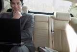 Foto: Businessman sitting in backseat of car, wearing mobile phone hands-free device, using laptop, smiling, front view, portrait (tilt) Businessman sitting in backseat of car, wearing mobile phone hands-free device, using laptop, smiling, front view, portrait (tilt)