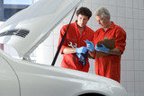 Photo: Two car mechanics, in red overalls and protective gloves, looking at car engine in auto repair shop, talking, mature man holding clipboard, side view Two car mechanics, in red overalls and protective gloves, looking at car engine in auto repair shop, talking, mature man holding clipboard, side view