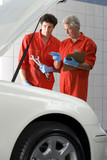 Photo: Two car mechanics, in red overalls and protective gloves, looking at car engine in auto repair shop, talking, mature man holding clipboard, colleague holding wrench, side view Two car mechanics, in red overalls and protective gloves, looking at car engine in auto repair shop, talking, mature man holding clipboard, colleague holding wrench, side view