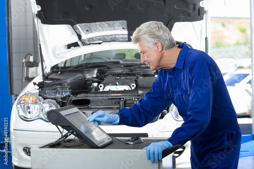 Mature car mechanic, in blue overalls, using computer in auto repair ...