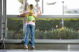 Photo: Girl (7-9), with ‘monkey’ rucksack, looking through large window in airport departure lounge, leaning against glass, watching aeroplane taking off, rear view Girl (7-9), with ‘monkey’ rucksack, looking through large window in airport departure lounge, leaning against glass, watching aeroplane taking off, rear view