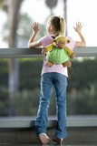 Photo: Girl (7-9), with ‘monkey’ rucksack, looking through large window in airport departure lounge, leaning against glass, rear view Girl (7-9), with ‘monkey’ rucksack, looking through large window in airport departure lounge, leaning against glass, rear view