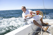 Photo: Mature man sitting on deck of yacht out at sea, leaning over side of boat, turning rope pulley, smiling Mature man sitting on deck of yacht out at sea, leaning over side of boat, turning rope pulley, smiling