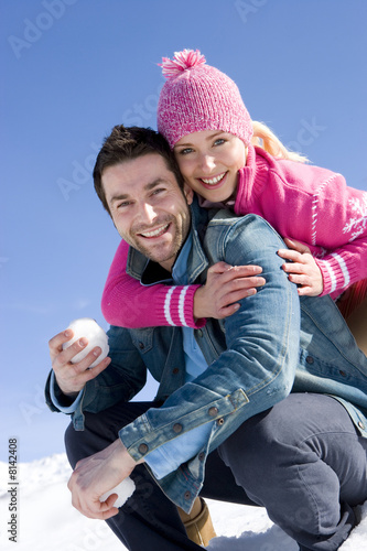 Photo: Young couple embracing in snow, man holding snow ball, smiling, portrait Young couple embracing in snow, man holding snow ball, smiling, portrait
