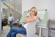 Photo: Woman sitting by blue prints on drafting board in home office, smiling portrait, man standing in background Woman sitting by blue prints on drafting board in home office, smiling portrait, man standing in background