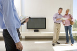 Photo: Male real estate agent and mature couple standing in living room, couple in background Male real estate agent and mature couple standing in living room, couple in background