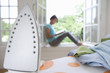 Iron on ironing board, woman on window sill reading in background, focus on iron