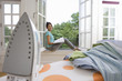 Woman with book on window sill, smiling, portrait, iron on ironing board in foreground (differential focus)