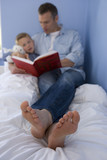 Photo: Father reading to son (4-6) in bed, close-up of father's feet (differential focus) Father reading to son (4-6) in bed, close-up of father's feet (differential focus)